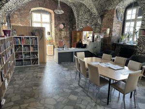 zdjęcie czytelni w Zamku w Leśnicy, na pierwszym planie stoi duży stół z krzesłami, w tle półki z ksiązkami i onieta z laptopem siedząca przy biurku