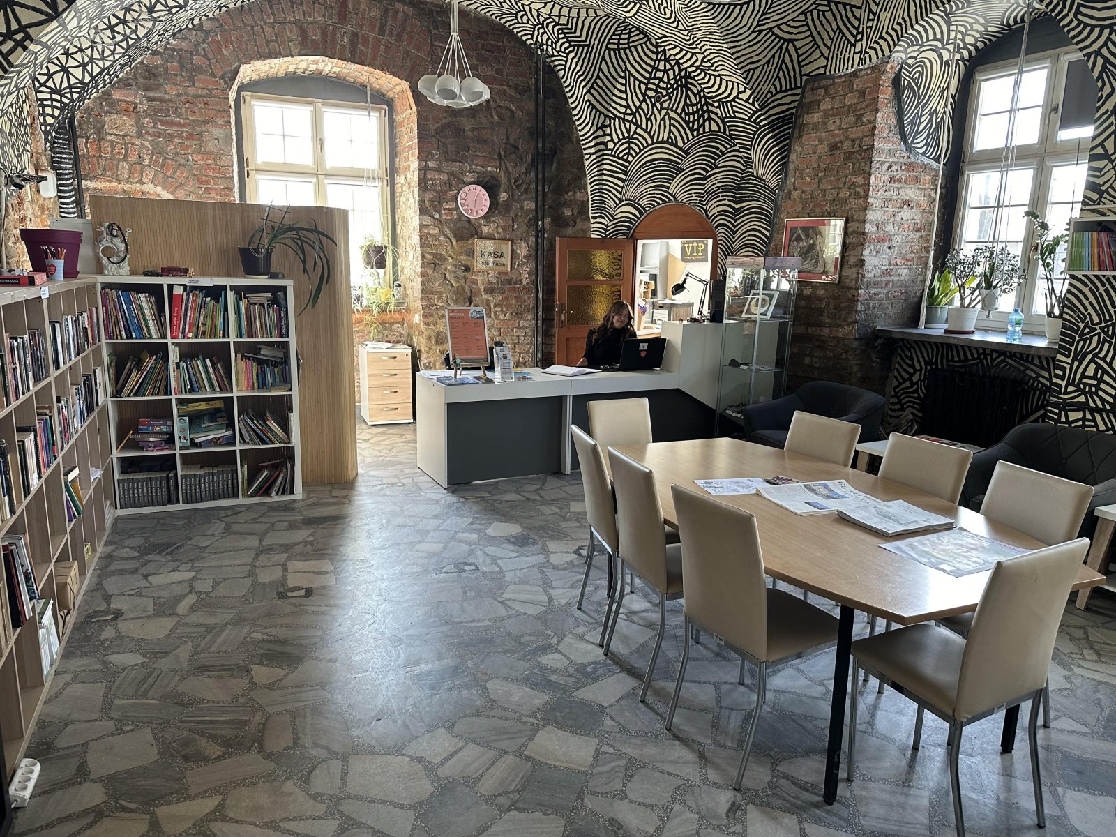 zdjęcie czytelni w Zamku w Leśnicy, na pierwszym planie stoi duży stół z krzesłami, w tle półki z ksiązkami i onieta z laptopem siedząca przy biurku